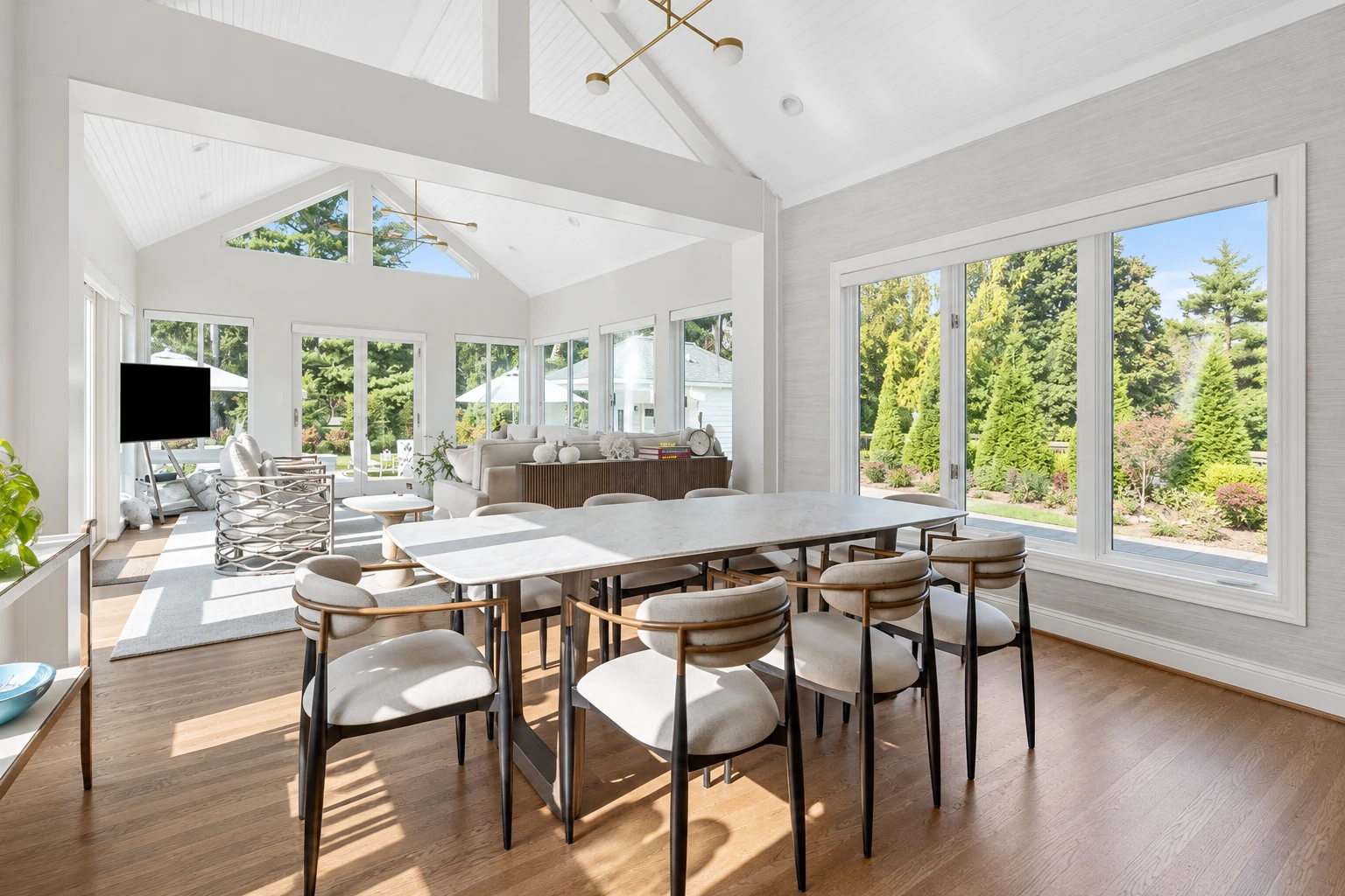 A bright sunroom addition features a vaulted white ceiling, large windows, and a dining table with upholstered chairs.