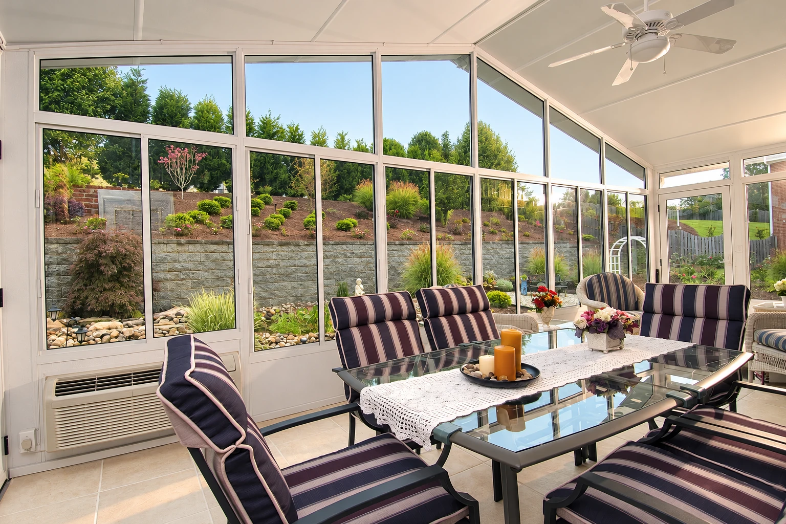 Bright sunroom with tall white-framed windows, striped lounge chairs, and a glass dining table overlooking a landscaped garden.