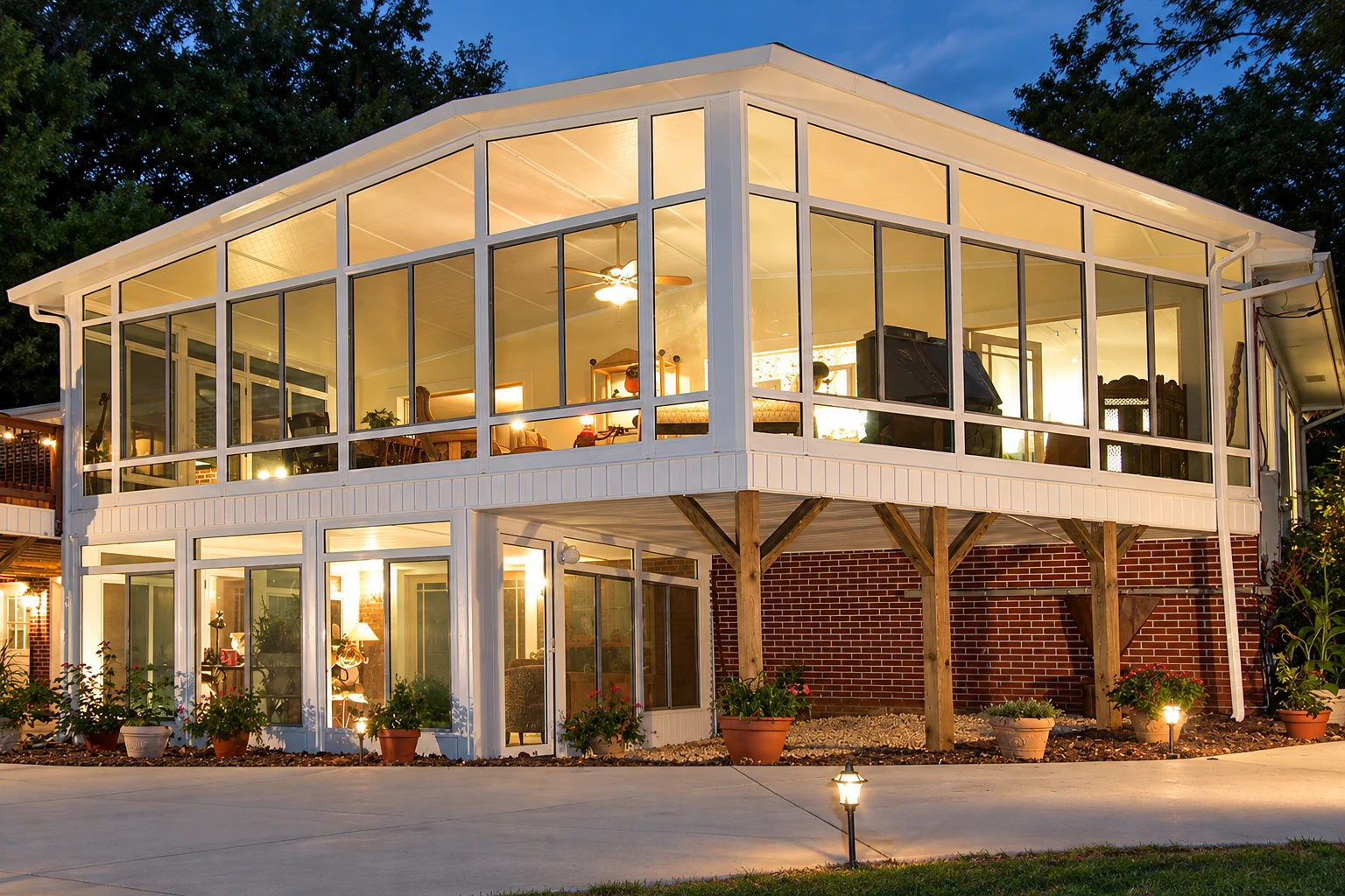 Two-story sunroom with white-framed glass walls, glowing warmly at dusk beside a brick house.